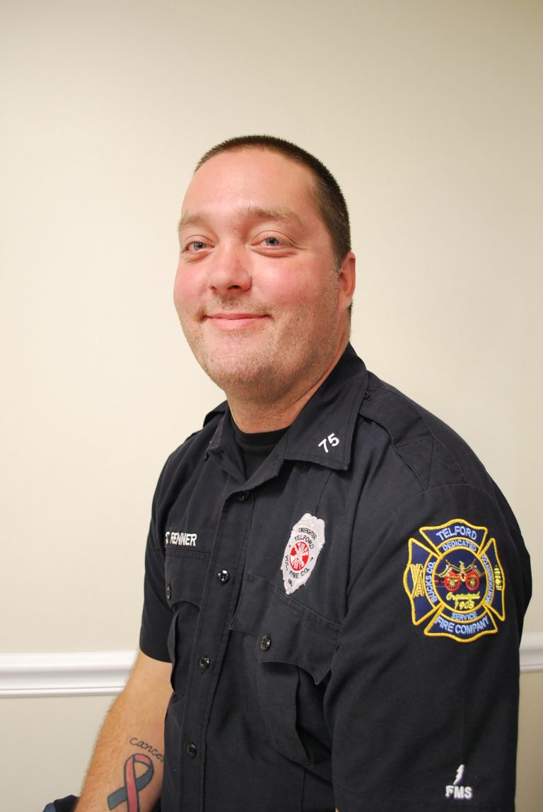 A firefighter in uniform with Telford Fire Company patches sits smiling against a plain light-colored wall, showing a tattoo on his forearm.