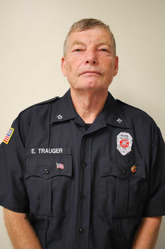 A man in a dark blue firefighter uniform stands against a plain light background. His shirt displays patches, a badge, and the name "E. Trauger." He has short hair and a neutral expression.