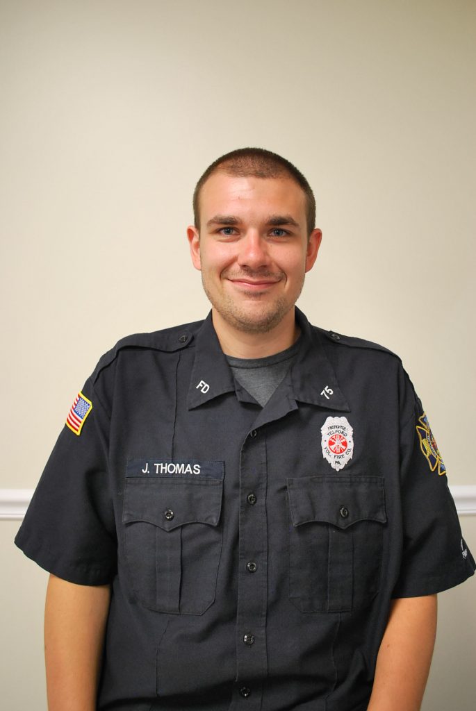 A man in a dark uniform with badges and the name "J. Thomas" smiles at the camera, standing against a plain beige wall.