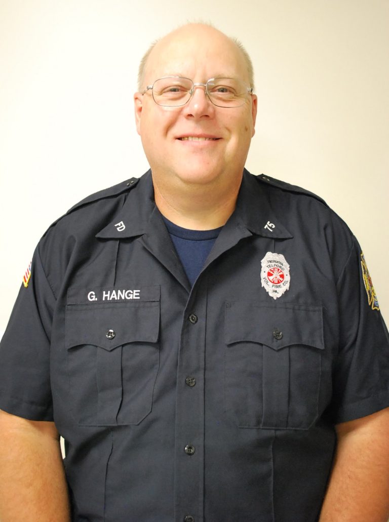 A smiling man wearing glasses and a dark blue fire department uniform with badges and a name tag that reads "G. HANGE," standing against a plain light background.