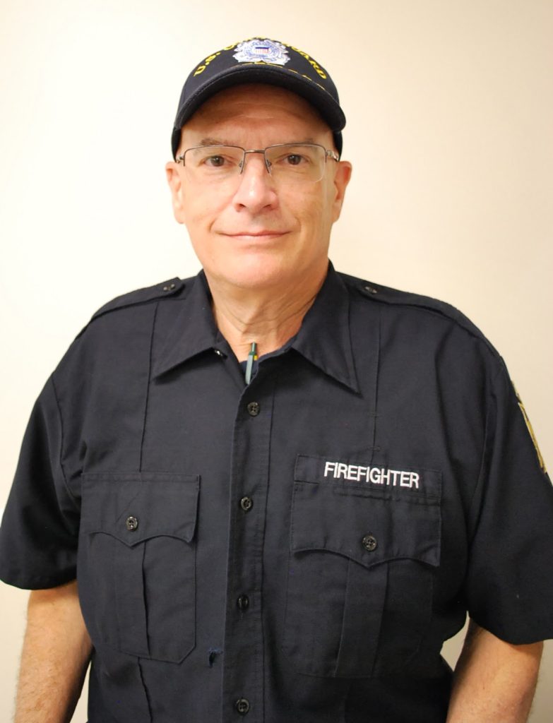 A middle-aged man wearing glasses, a dark blue uniform shirt labeled "FIREFIGHTER," and a cap, standing against a plain light-colored background.