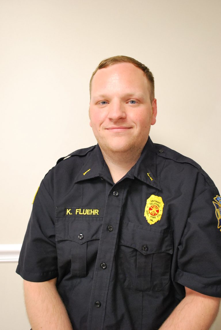 A man in a black uniform with fire department badges and the name "K. Fluehr" on his shirt stands in front of a plain light-colored wall, smiling slightly at the camera.