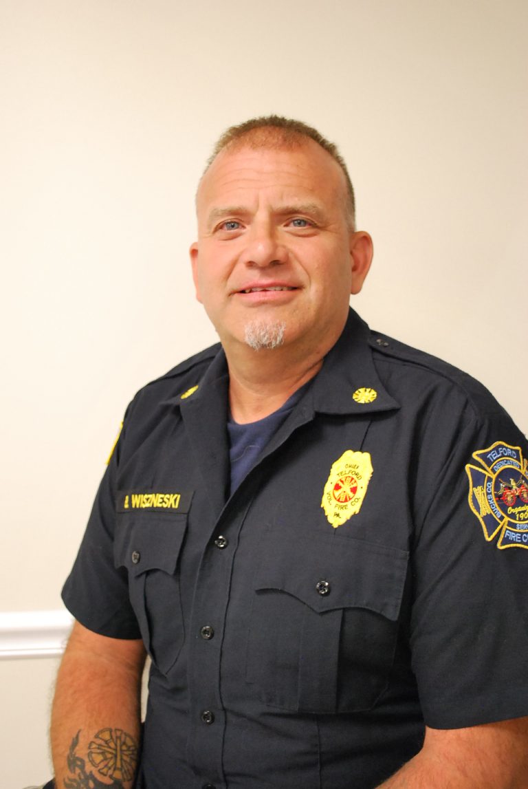 A man in a dark fire department uniform with badges sits and smiles at the camera against a plain, light-colored background. He has short hair, a goatee, and a tattoo on his left forearm.