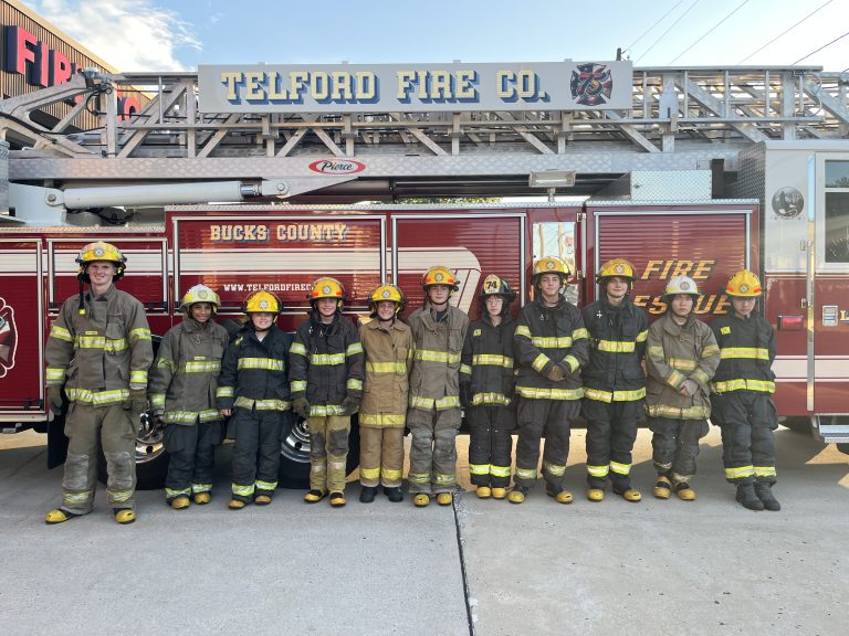 Eleven firefighters in uniform and helmets stand in a line in front of a red fire truck labeled “Telford Fire Co.,” smiling for a group photo outside the station.