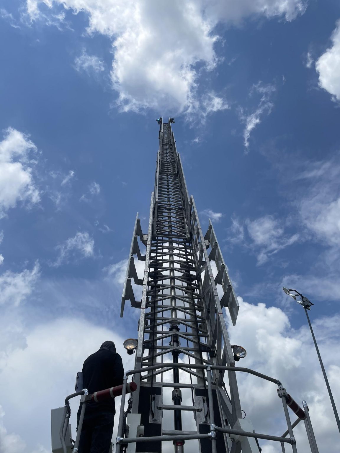 A person stands at the base of an extended metal ladder reaching high into a partly cloudy sky, with antennas or platforms attached along its sides.