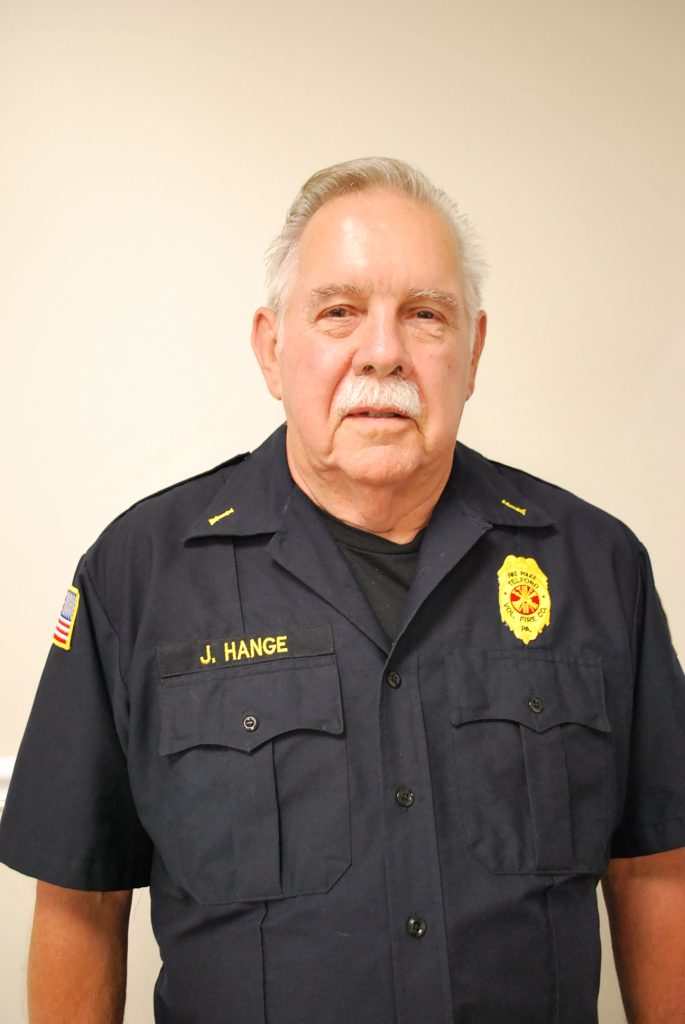 An older man with gray hair and a mustache wearing a dark uniform shirt with a fire department badge patch and a name tag reading "J. Hange" stands against a plain light-colored background.