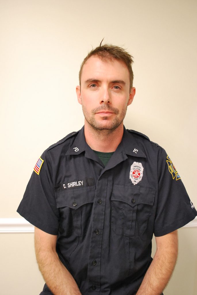 A man in a navy blue firefighter uniform sits against a beige wall. His shirt displays badges, an American flag patch, and a name tag that reads "C. Shirley." He has short hair and a neutral expression.