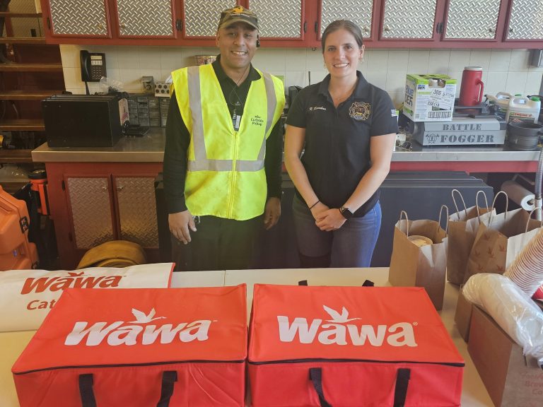 Two people, one in a yellow safety vest and one in a navy shirt, stand behind a table with red Wawa catering bags and paper bags, inside a room with kitchen equipment and supplies.
