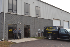 Two people stand outside the entrance of a gray industrial building with large windows. A black company truck with yellow lettering is parked nearby, and two large garage doors are visible in the background.