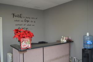 A small reception area with a gray wall featuring a quote about faith, a desk with a red poinsettia plant and holiday card, a water cooler, and some decorative items.