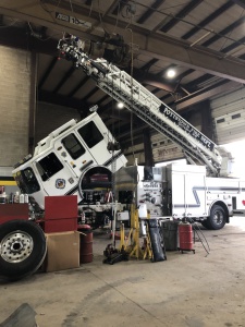 A fire truck with its cab tilted forward and ladder extended is undergoing maintenance inside a fire station garage. Various tools and equipment are scattered around the vehicle.