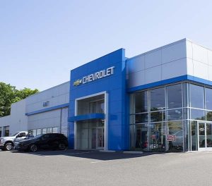 A modern Chevrolet car dealership building with blue and white exterior, large windows, and several cars parked outside on a sunny day.