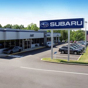 A Subaru car dealership with a large Subaru sign, several parked cars in front of the showroom, and trees in the background under a clear blue sky.