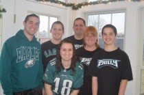 Six people stand together indoors, smiling at the camera. They are wearing Philadelphia Eagles shirts and jerseys, with a window and holiday garland visible in the background.