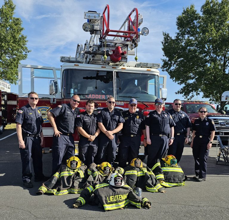 Nine firefighters in uniform stand in front of a fire truck labeled "Ladder 75." Four sets of firefighting gear and helmets are arranged on the ground in front of them. Trees and vehicles are visible in the background.