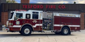 A red and white fire truck labeled "Telford Fire Co." is parked in front of a fire station with matching "Telford Fire Co." signage. The truck displays various gauges and equipment compartments.