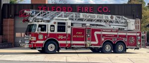 A red fire truck with a ladder is parked in front of Telford Fire Company’s building, which has large garage doors and a sign reading "TELFORD FIRE CO." The truck is marked "Montgomery County Fire Rescue.