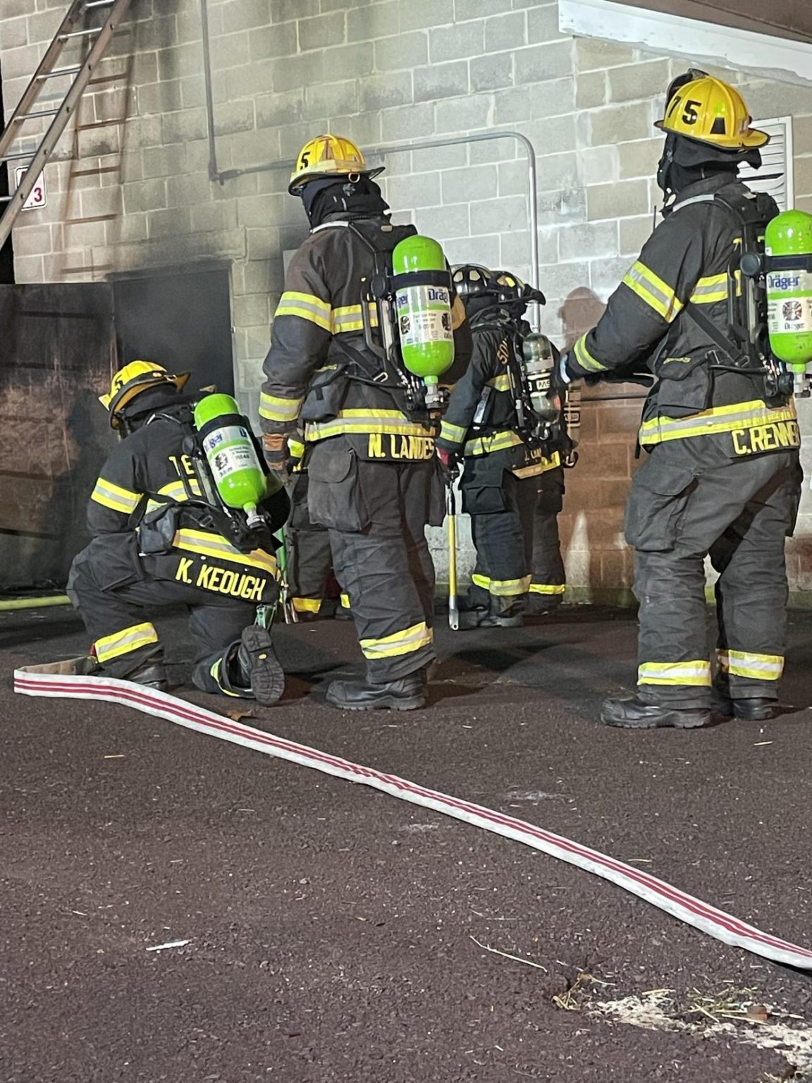 Four firefighters in full gear with yellow helmets and green oxygen tanks work together near a building. Two stand while two crouch by a wall. A fire hose lies on the pavement nearby.