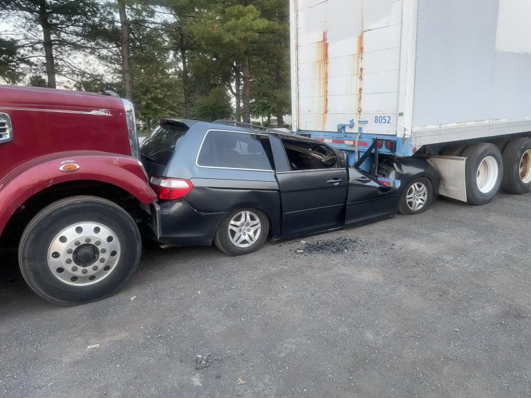 A black car is crushed between the front of a red truck and the rear of a white semi-trailer in a parking lot, with visible damage to the car's roof and sides. Trees are visible in the background.