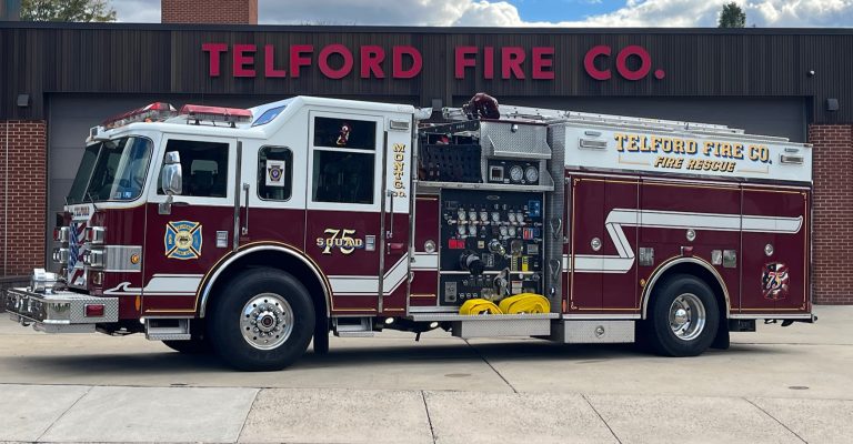 A maroon and white fire truck labeled "Telford Fire Co. Fire Rescue" is parked in front of a building with a matching sign. The truck has various equipment and hoses visible in its side compartments.