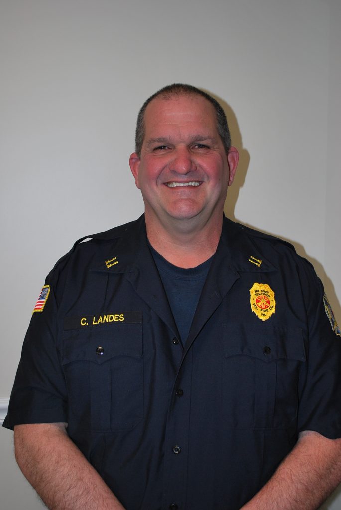 A smiling man in a dark police uniform stands against a plain light-colored wall. The uniform displays a badge, shoulder patches, and a name tag reading "C. Landes.