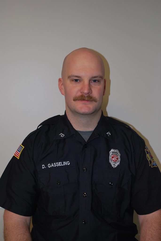 A man in a black uniform with a badge and name tag reading "D. Gasseling" sits against a plain background. He has a bald head, a mustache, and an American flag patch on his sleeve.