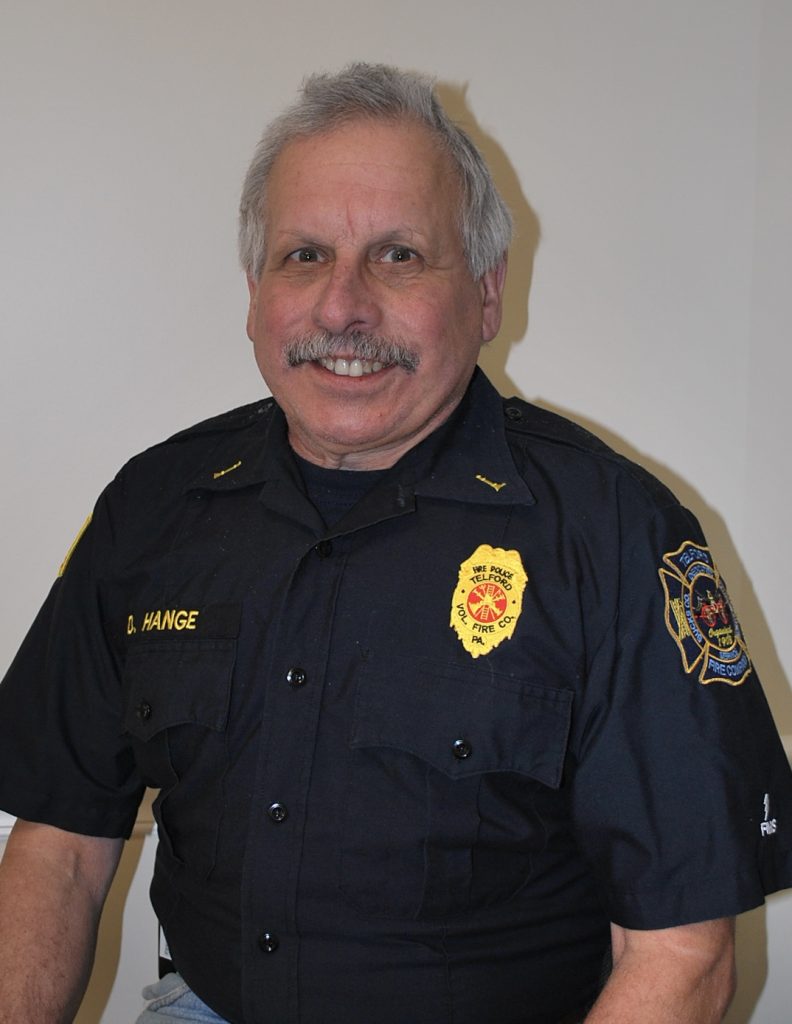 A smiling older man with gray hair and a mustache wearing a black fire department uniform sits against a plain light-colored wall.