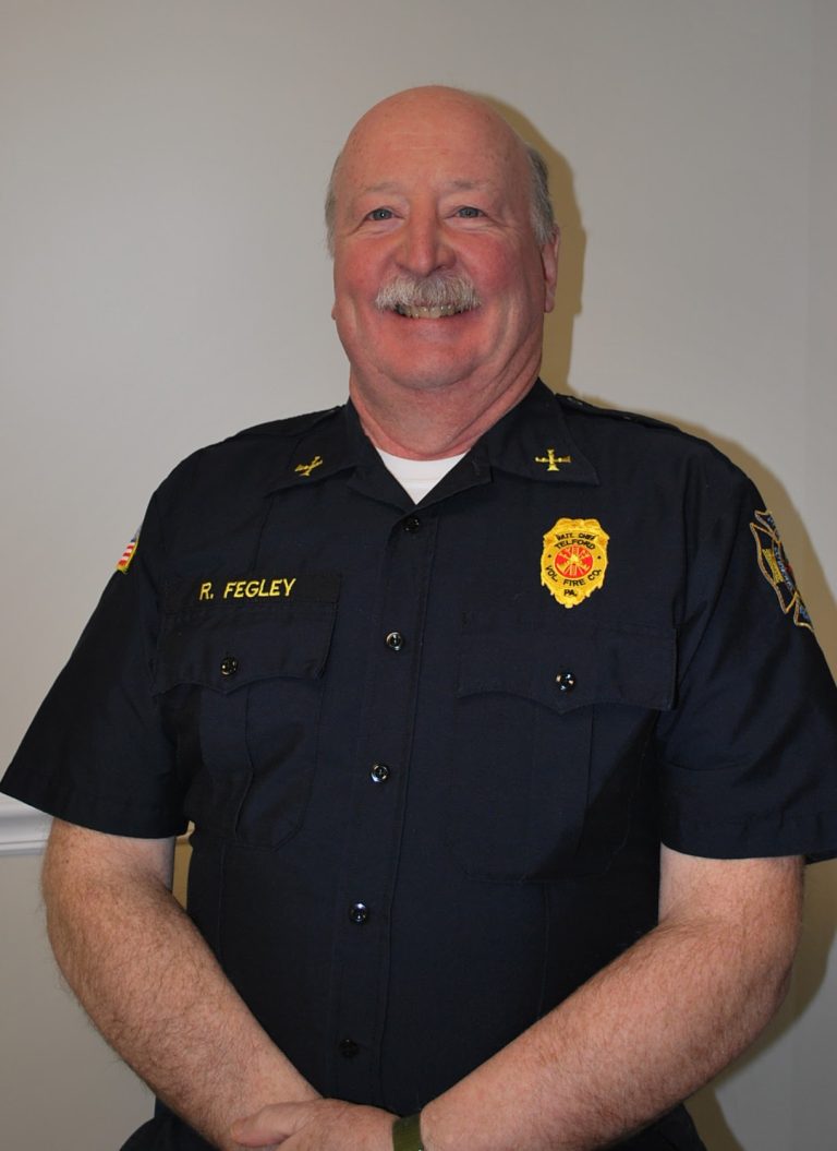 A smiling man in a dark uniform with badge and name tag "R. Fegley" stands against a plain light-colored wall. He has short, light hair, a mustache, and his hands are clasped in front of him.