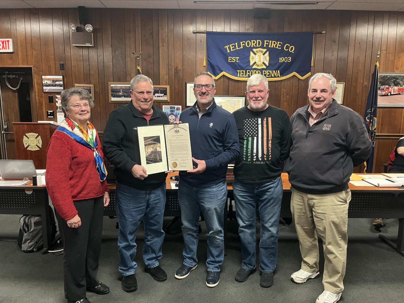 Five adults stand together smiling in a wood-paneled room at Telford Fire Company; one man holds a framed photo and a proclamation. A Telford Fire Co. banner and a meeting table are visible in the background.