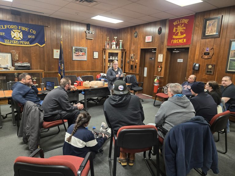 A group of people sits in a circle listening to a woman speaking in a wood-paneled room decorated with fire company banners, trophies, and firefighter-themed items.