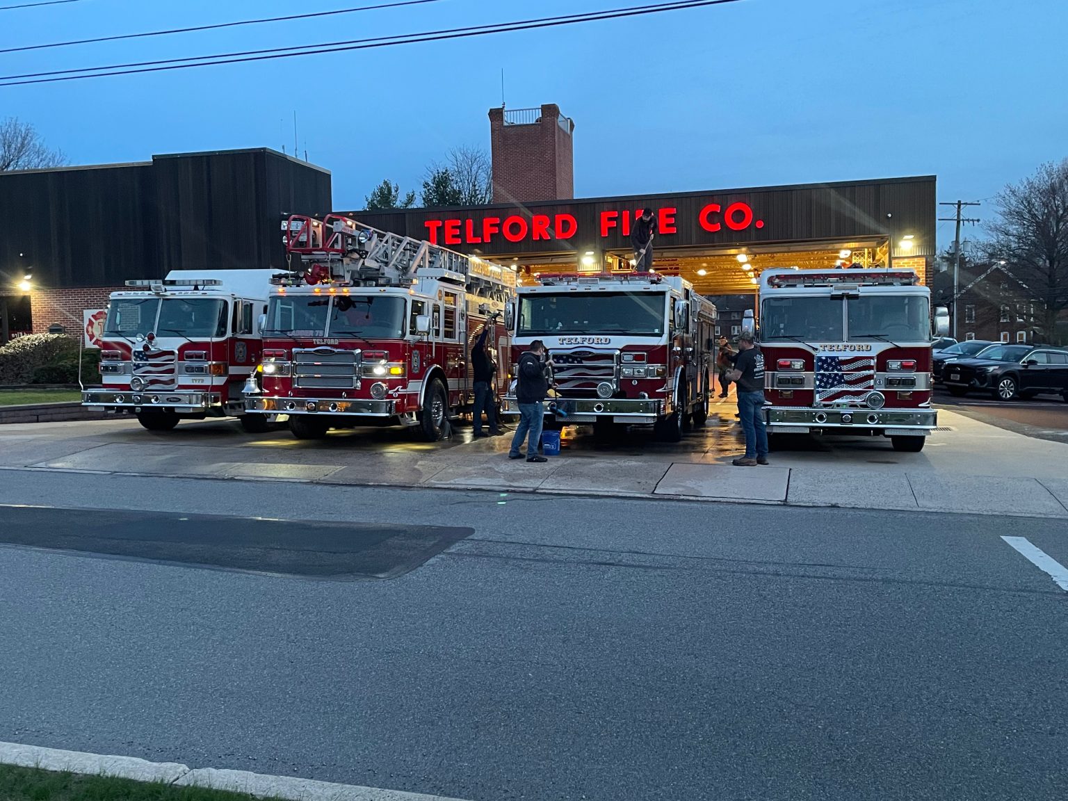 Four fire trucks are parked in front of the Telford Fire Company station at dusk, with several firefighters standing nearby. The station's illuminated sign and the trucks' reflective stripes are visible.