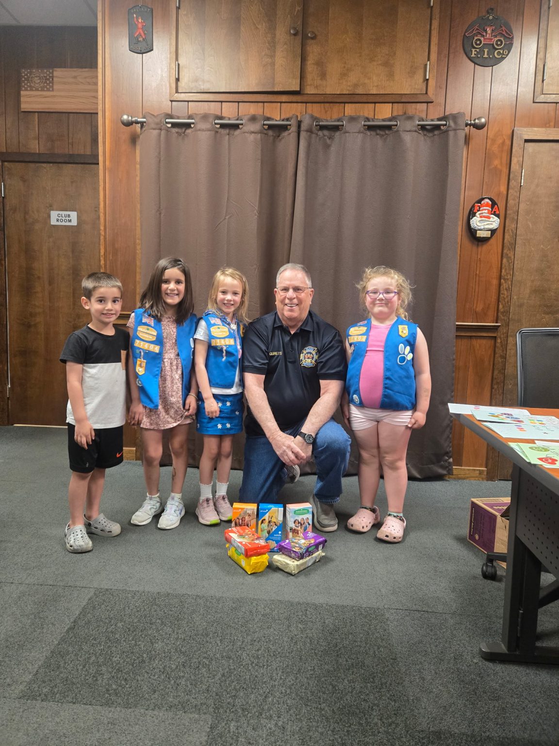 Five smiling children, four wearing blue vests with patches, stand around an older man kneeling in the center. In front of them are assorted snacks and food items. They are indoors, with wooden walls in the background.