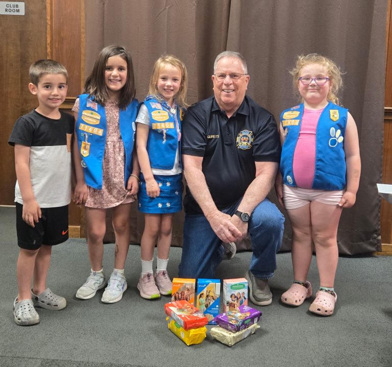 Five smiling children, four wearing blue vests with patches, stand around an older man kneeling in the center. In front of them are assorted snacks and food items. They are indoors, with wooden walls in the background.