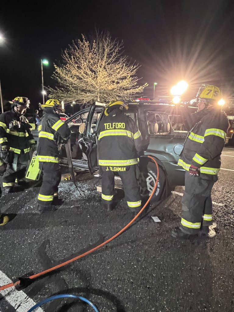 Four firefighters in full gear work at night to rescue someone from a damaged car with shattered glass, using equipment and hoses. Bright lights and a tree are visible in the background.