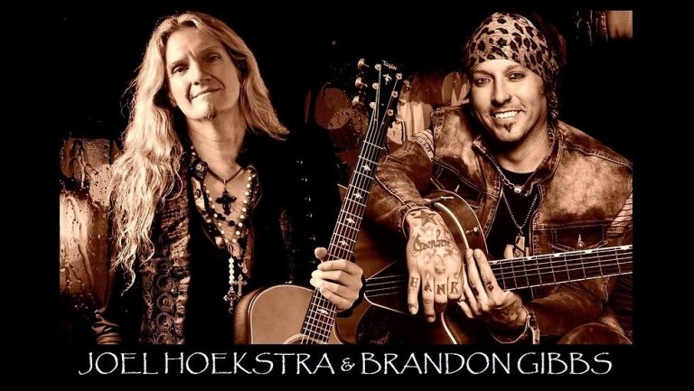 Two musicians pose with acoustic guitars. The musician on the left has long blonde hair and wears layered necklaces; the one on the right wears a patterned headscarf and a leather jacket. Text reads: "Joel Hoekstra & Brandon Gibbs.
