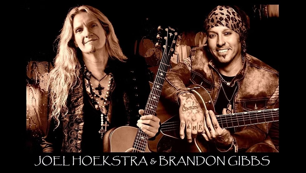 Two musicians pose with acoustic guitars. The musician on the left has long blonde hair and wears layered necklaces; the one on the right wears a patterned headscarf and a leather jacket. Text reads: "Joel Hoekstra & Brandon Gibbs.