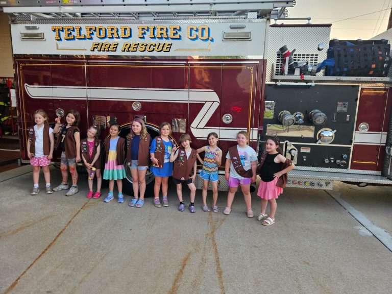 A group of young Girl Scouts wearing sashes and vests stand in a line in front of a Telford Fire Company fire truck.