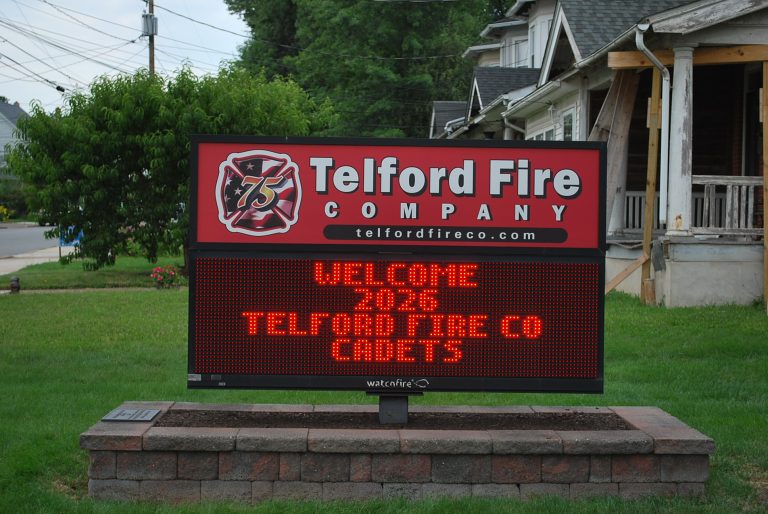 A digital sign outside Telford Fire Company reads, “WELCOME 2023 TELFORD FIRE CO CADETS.” The sign is red with a fire company logo at the top. Nearby houses and green grass are visible in the background.