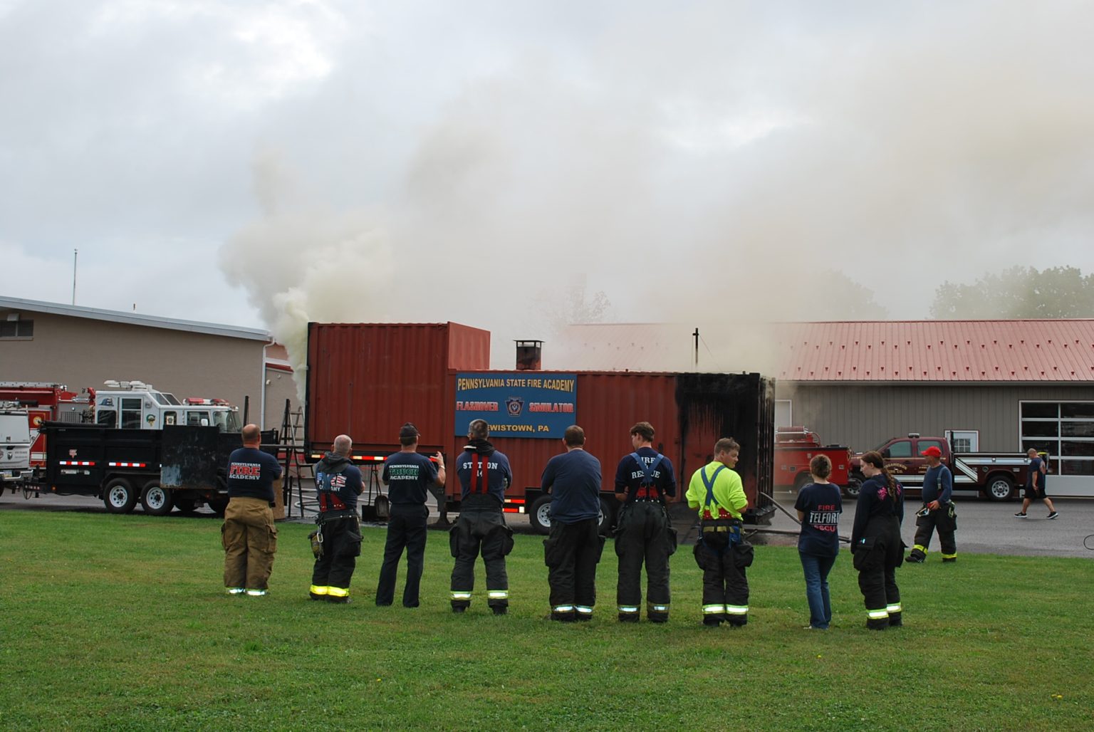 A group of firefighters stand on grass, watching smoke rise from a training building labeled “Fire Academy.” Fire trucks and equipment are visible in the background.